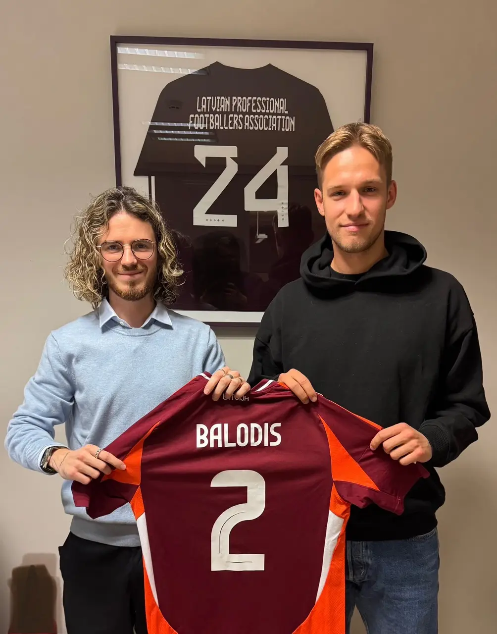 Marks Amosejevs and Daniels Balodis holding a Latvia shirt with the name Balodis and number 2
